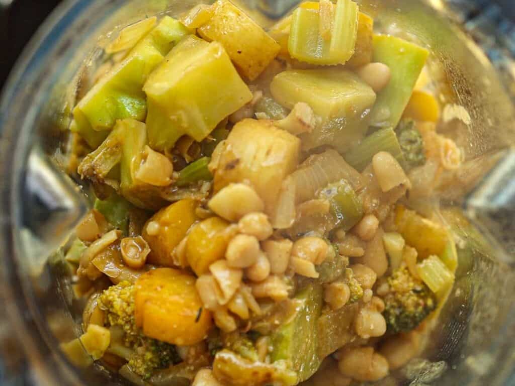 Close-up of a blender containing chopped vegetables, including broccoli, carrots, and onion, mixed with white beans and seasonings. The ingredients are layered in a glass container, suggesting preparation for blending.